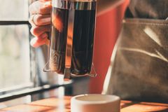 A barista uses a French press to brew coffee in a well-lit cafe.