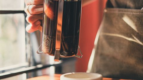 A barista uses a French press to brew coffee in a well-lit cafe.