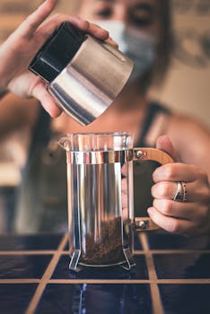 Barista adding coffee grounds to a French press in a café setting.