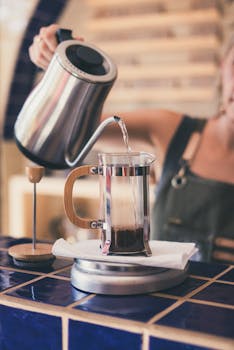 A barista pouring hot water into a French press in a cozy café setting.