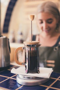 A French press coffee maker in a warm, inviting cafe setting with a smiling barista in the background.
