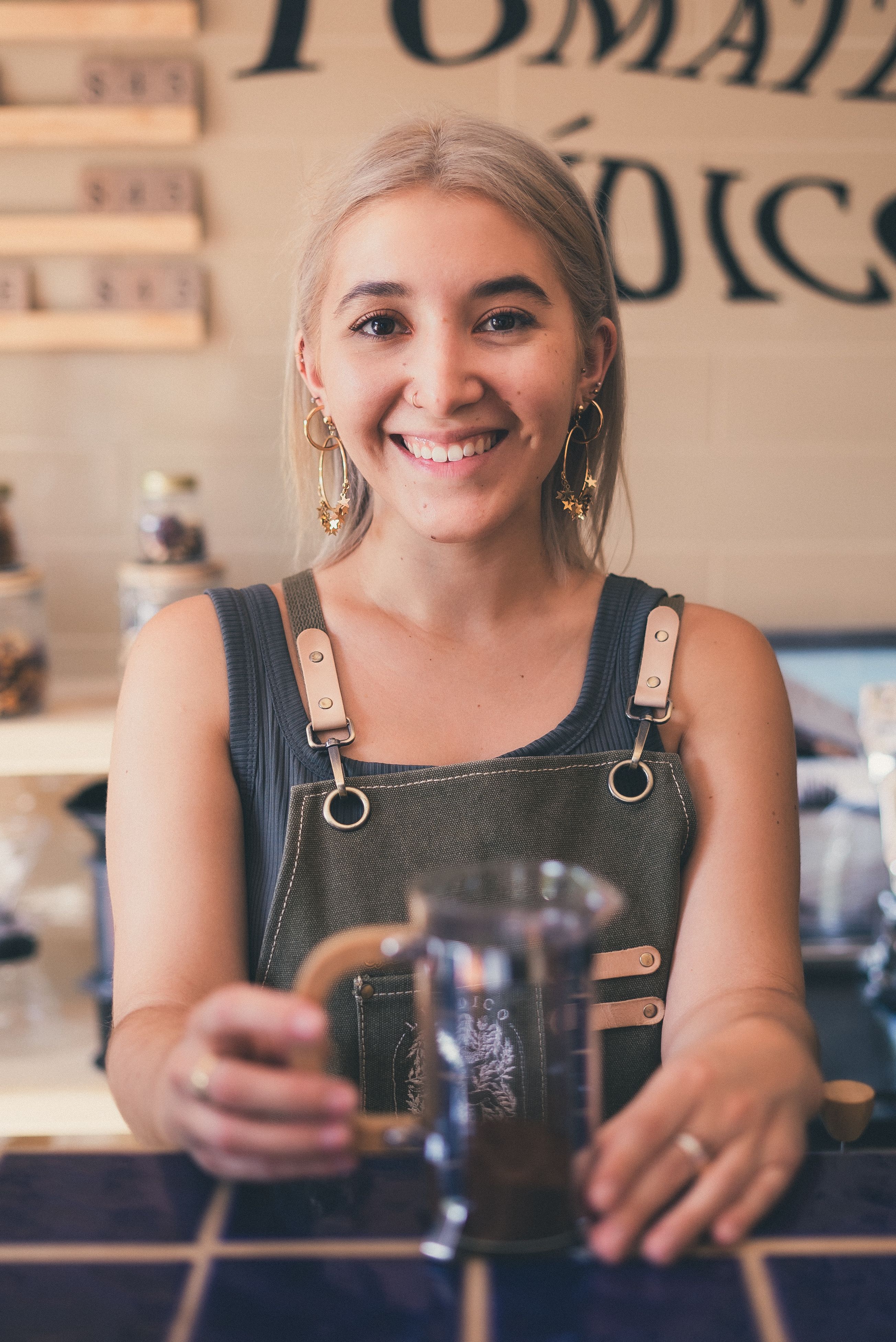 A stylish smiling barista holding a French press in a cozy coffee shop.