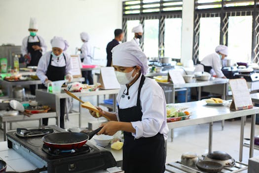 Chefs practicing culinary skills in a training kitchen setting, wearing masks for safety.