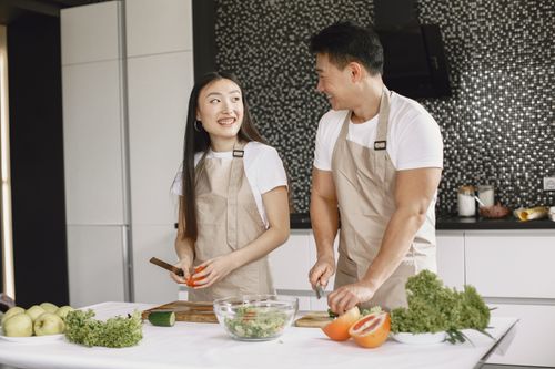 Free Asian couple joyfully preparing food together indoors wearing matching aprons. Stock Photo