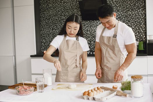Asian couple preparing food together in a modern kitchen, wearing casual aprons and smiling.
