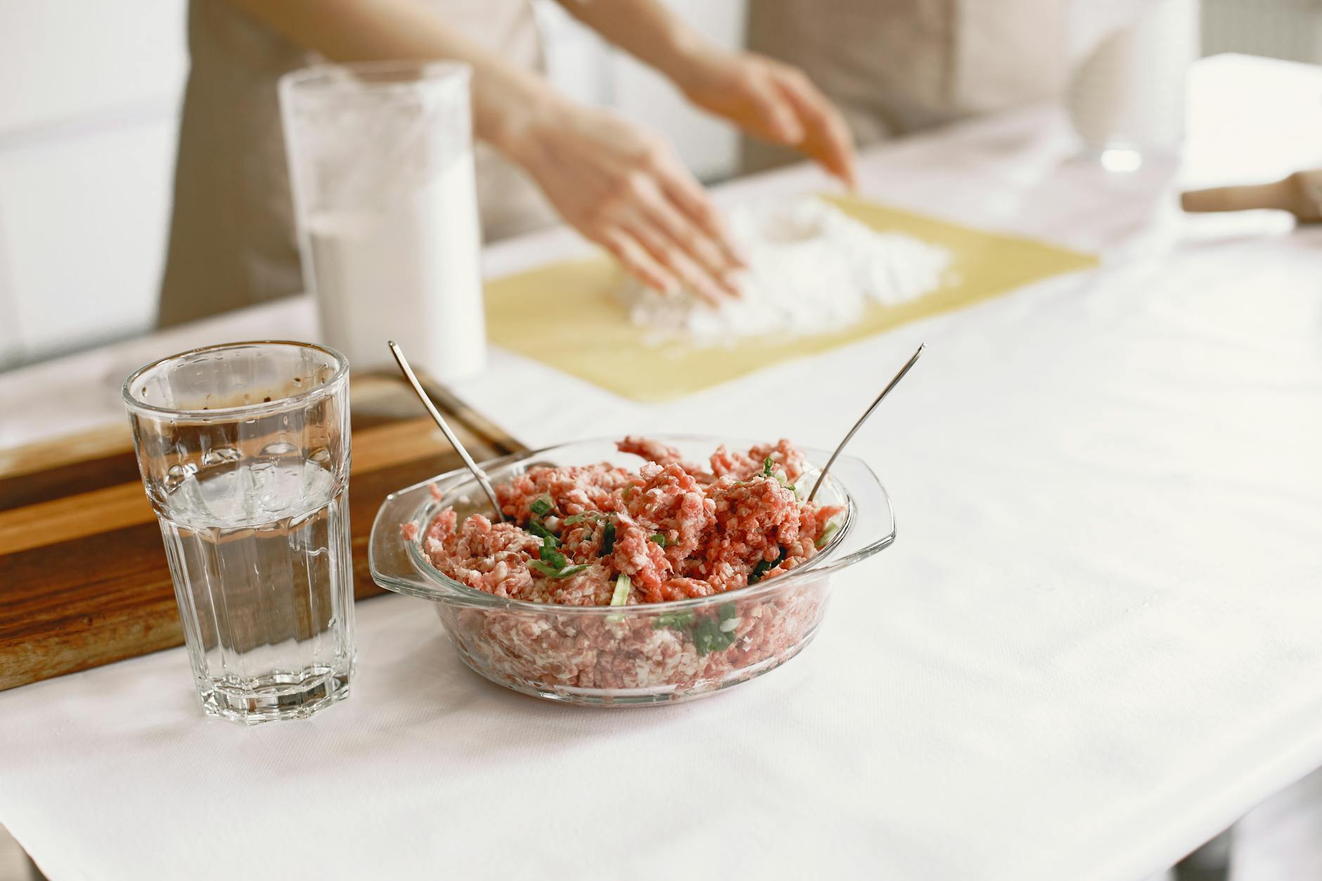 Raw ground meat in a glass bowl with ingredients being prepared nearby.