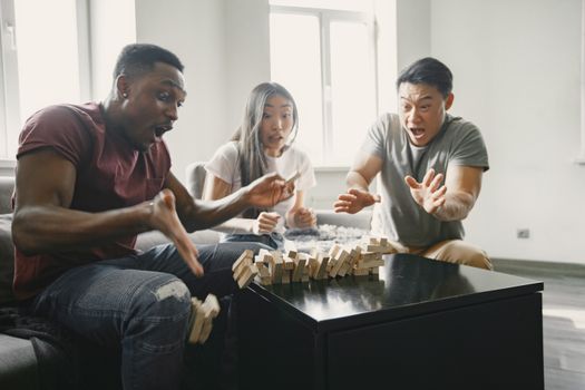 Three young adults intensely playing a game of Jenga indoors, capturing the moment of emotional reaction.