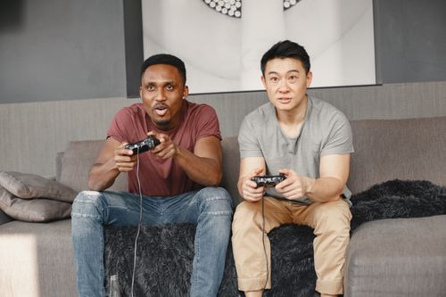 Free Two men sitting on a sofa, engaged in playing video games with controllers, showing excitement and concentration. Stock Photo