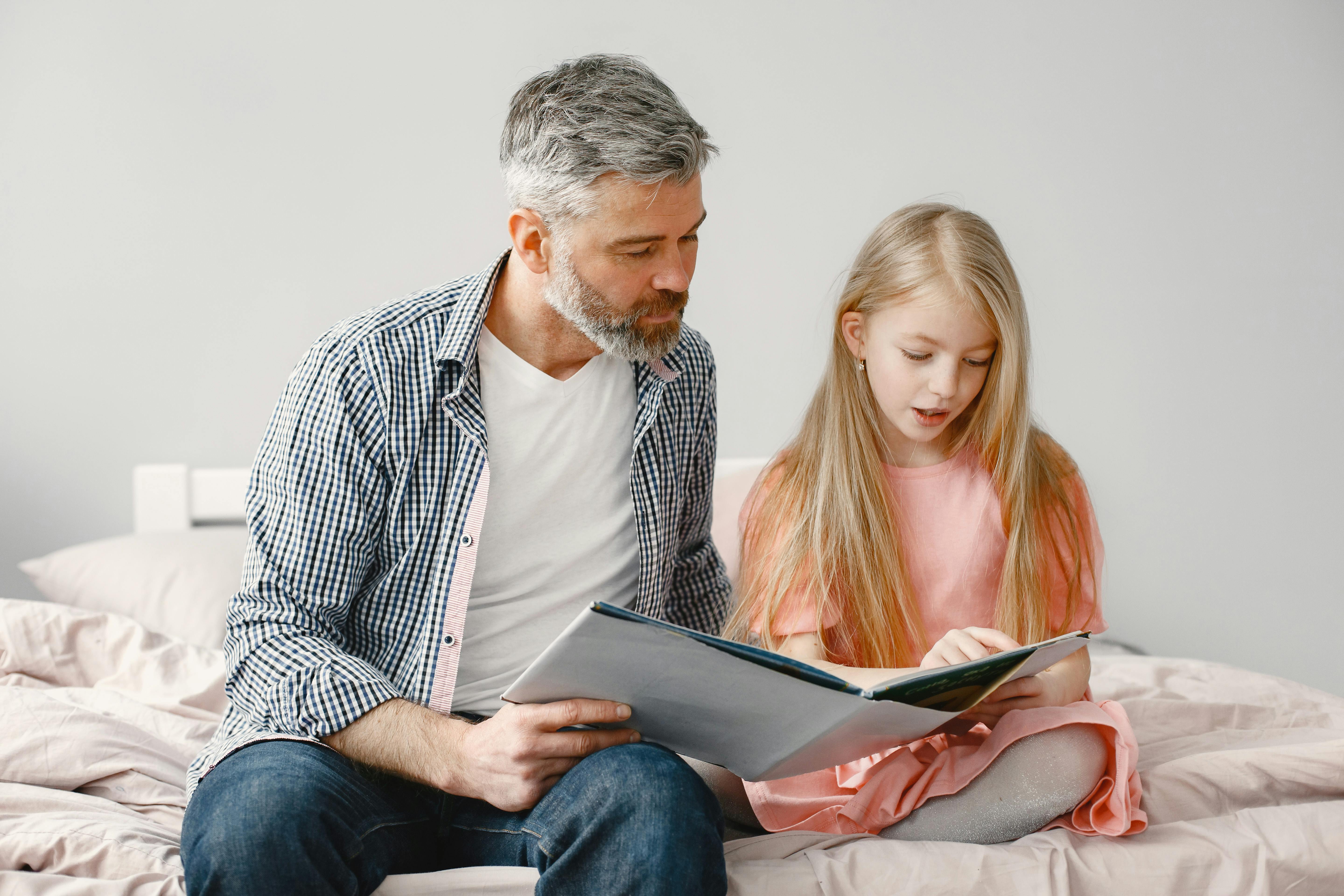 A father and daughter reading a book together on a bed in a cozy home setting.