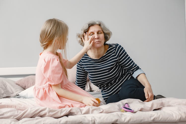 A Young Girl Putting An Under Eye Mask On Her Grandma's Face While Sitting On The Bed