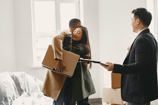 A happy couple hugs inside their new home with a realtor present, surrounded by moving boxes.