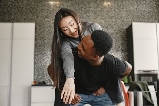 Happy African American man carrying Asian woman in playful embrace inside modern home.