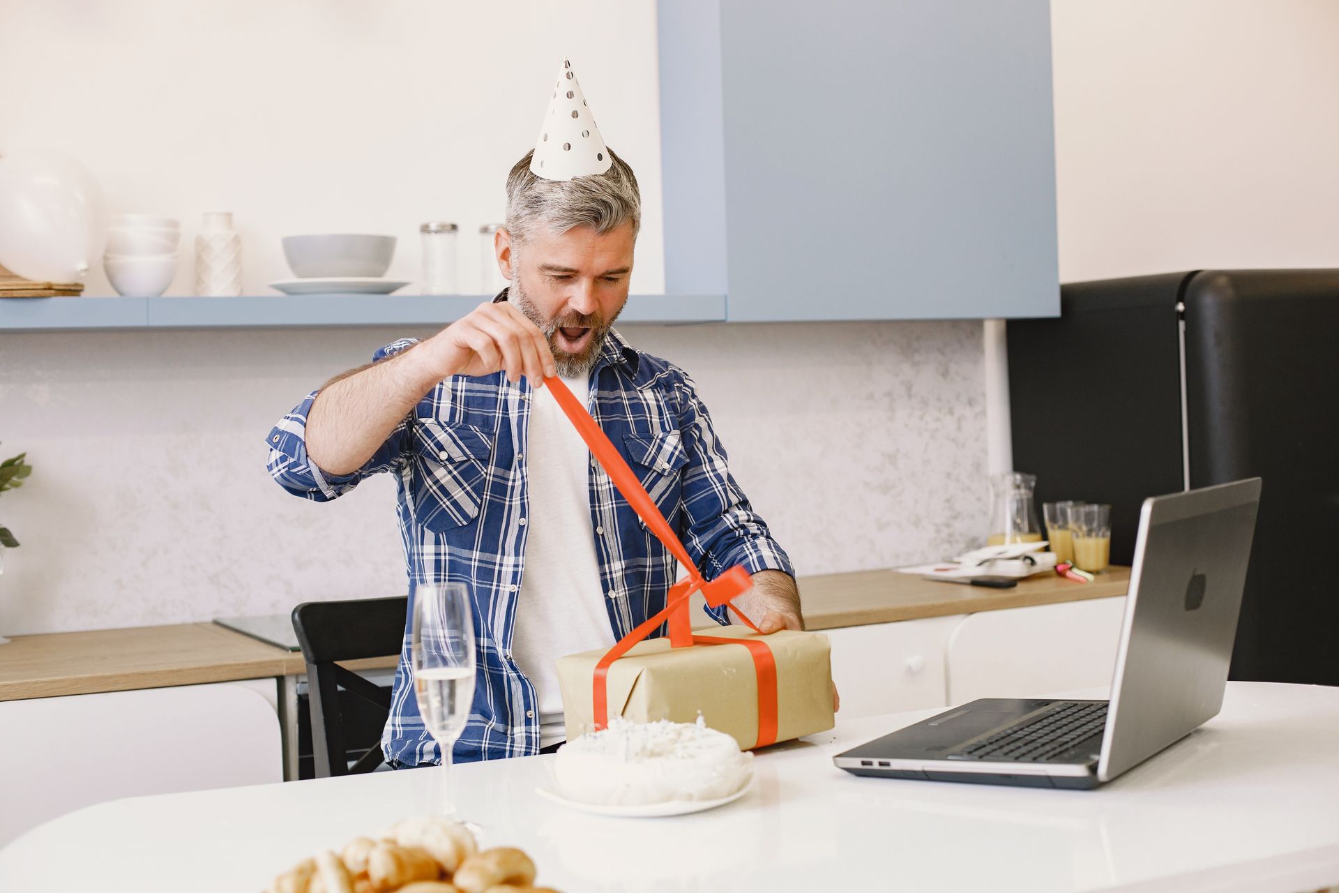 Happy man in party hat opening a gift during an online birthday celebration.