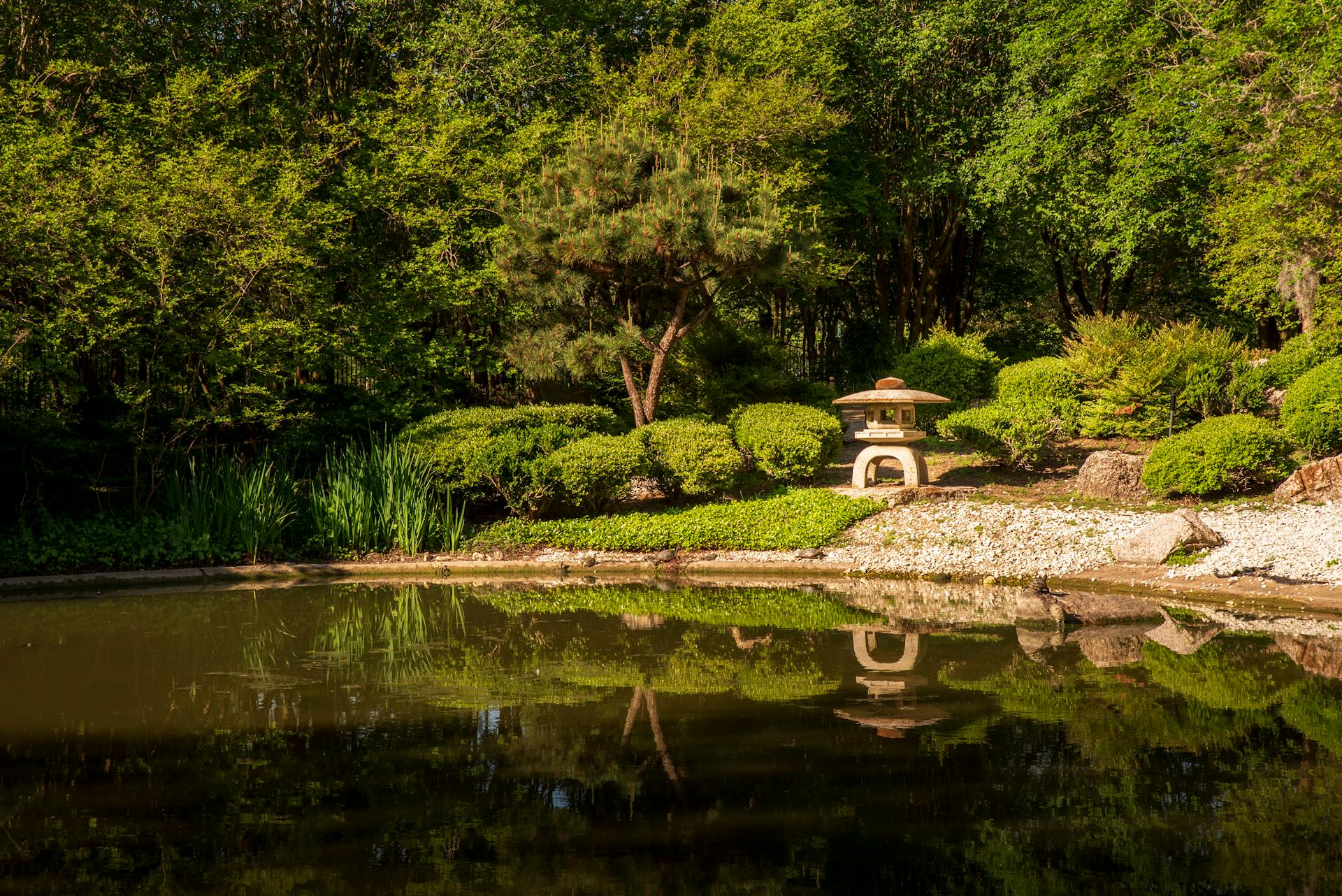 Tranquil Japanese garden scene with a pagoda-style stone lantern, lush greenery, and peaceful pond reflection.