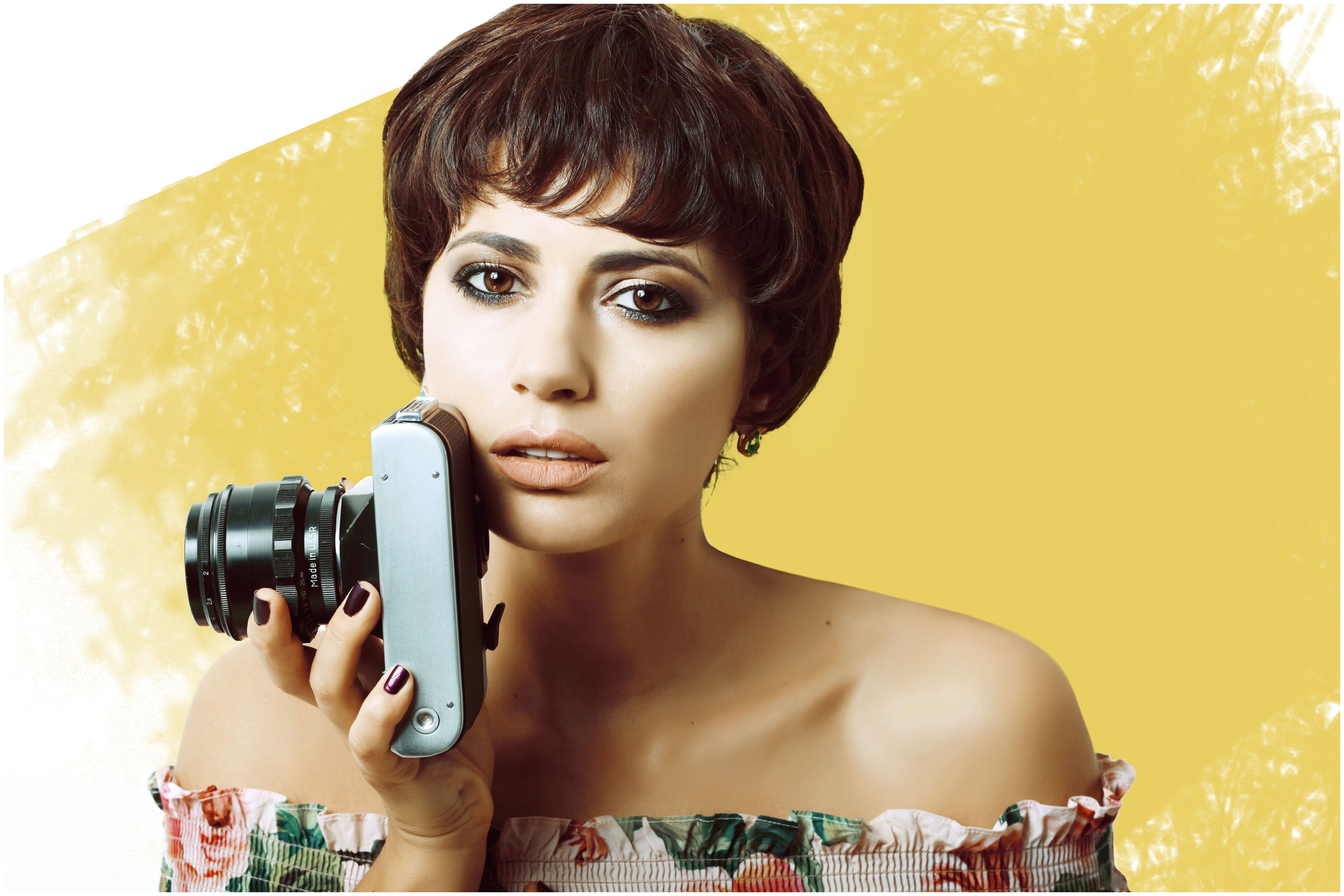 Charming female with makeup looking at camera while standing with professional photo camera on yellow painted background in light studio