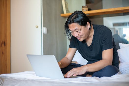 Man sits comfortably on his bed working remotely with a laptop, exemplifying modern freelance lifestyle.
