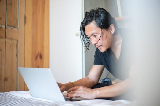 An Asian man focused on remote work using a laptop inside a cozy home setting.