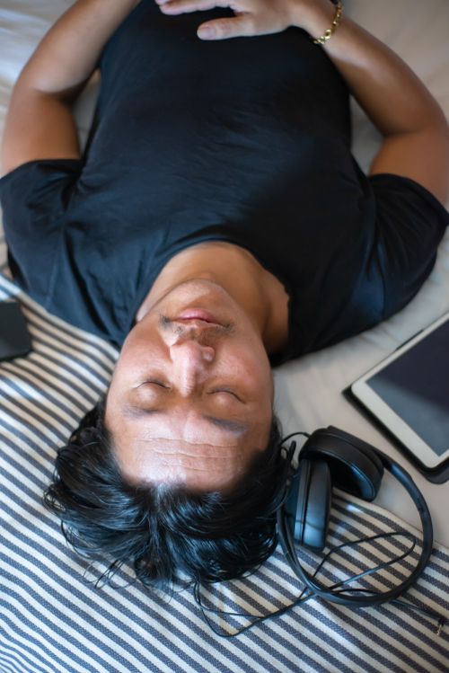 Free Man lying on bed with eyes closed, headphones, and tablet nearby, conveying relaxation. Stock Photo