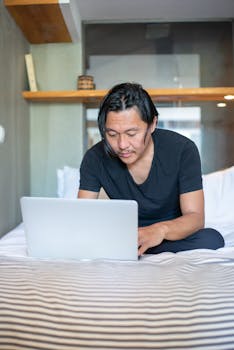 An Asian man using a laptop while sitting on a bed in a cozy home environment.