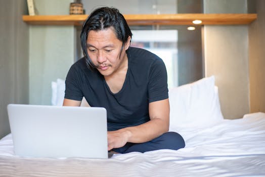 Asian man working remotely on a laptop in a cozy bedroom setting, exemplifying modern work-from-home lifestyle.