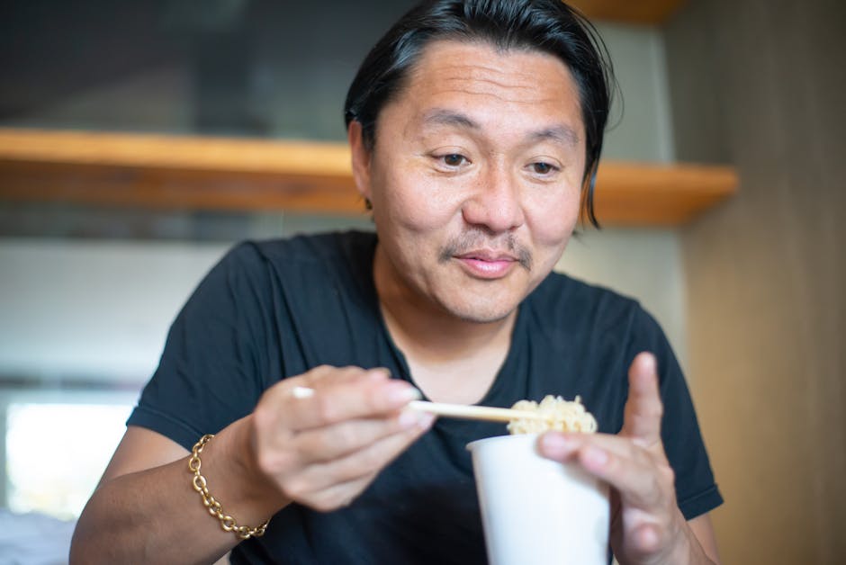 A man joyfully eating noodles with chopsticks from a cup indoors.