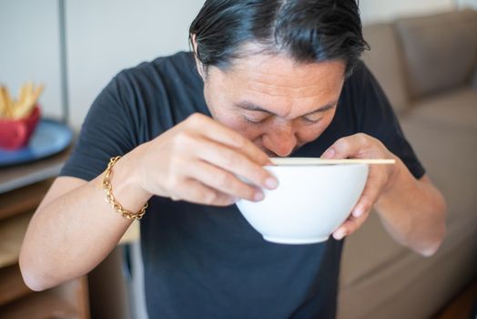 Asian man savoring noodles in a ceramic bowl, enjoying a peaceful meal at home with chopsticks.