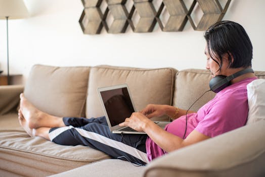 An Asian man in a pink shirt working on his laptop while wearing headphones, relaxing on a couch indoors.