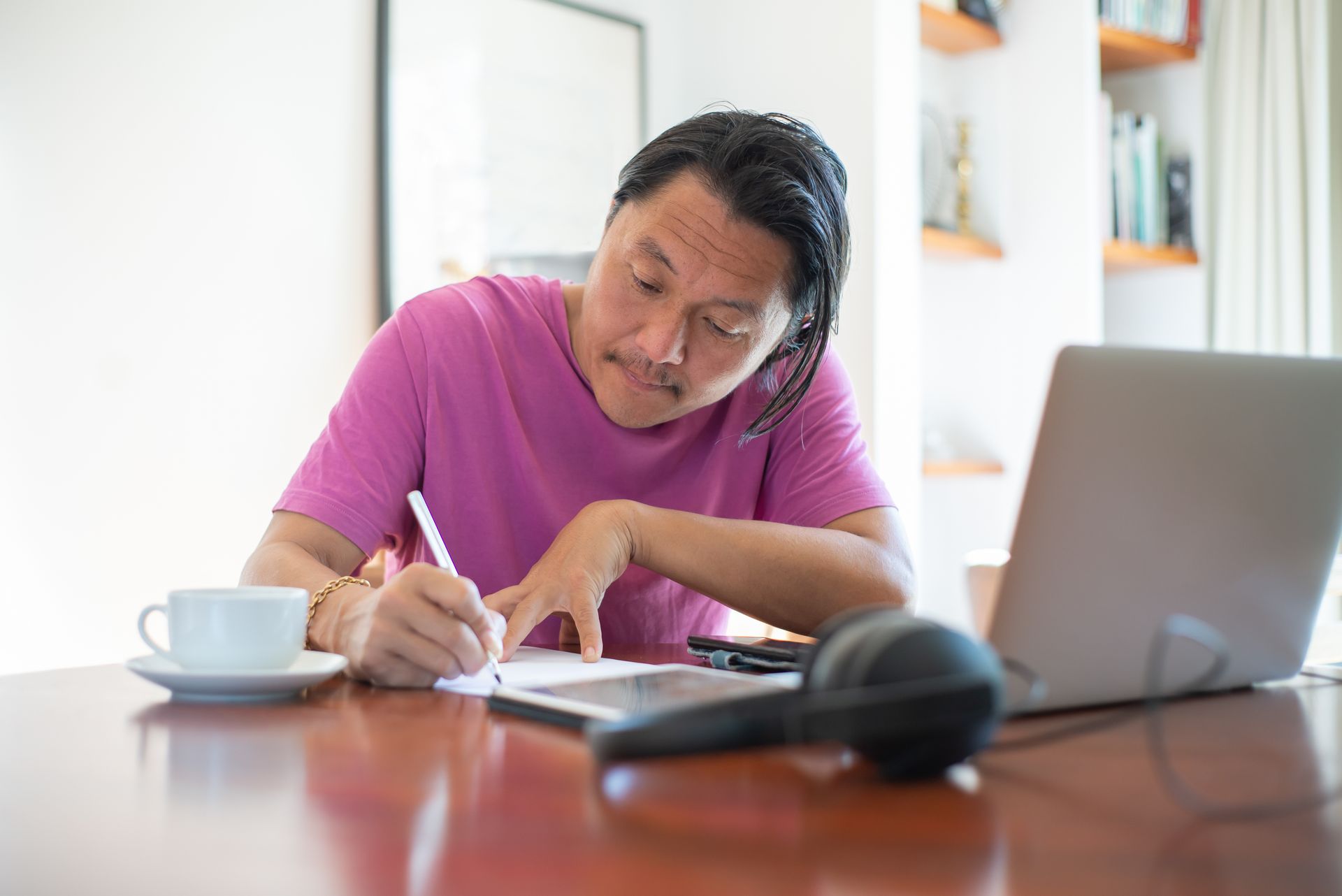 An adult man in a pink shirt works intently at a home office desk with a laptop and notebook.