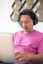 A Man in Pink Shirt Wearing Headphones while Looking at His Laptop