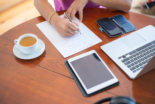 Close-up of a workspace with a laptop, tablet, smartphone, cup of tea, and a person writing notes.