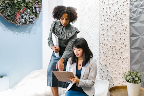 Free A Woman in White Blazer Sitting while Talking to the Woman Standing Beside Her Stock Photo