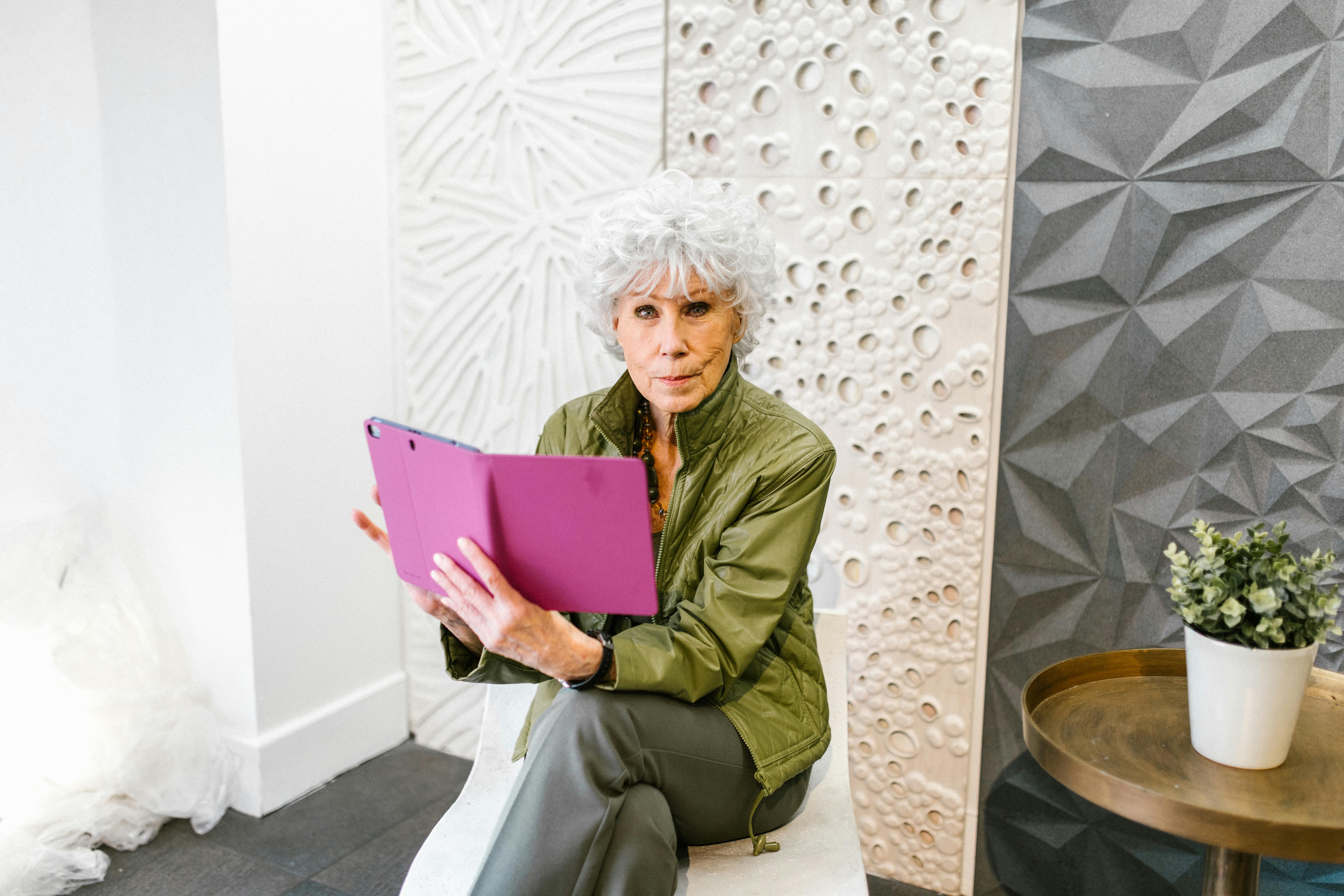 Elderly woman in a green jacket using a tablet in a stylish indoor setting.