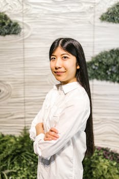 A confident woman in a white shirt stands with arms crossed indoors.