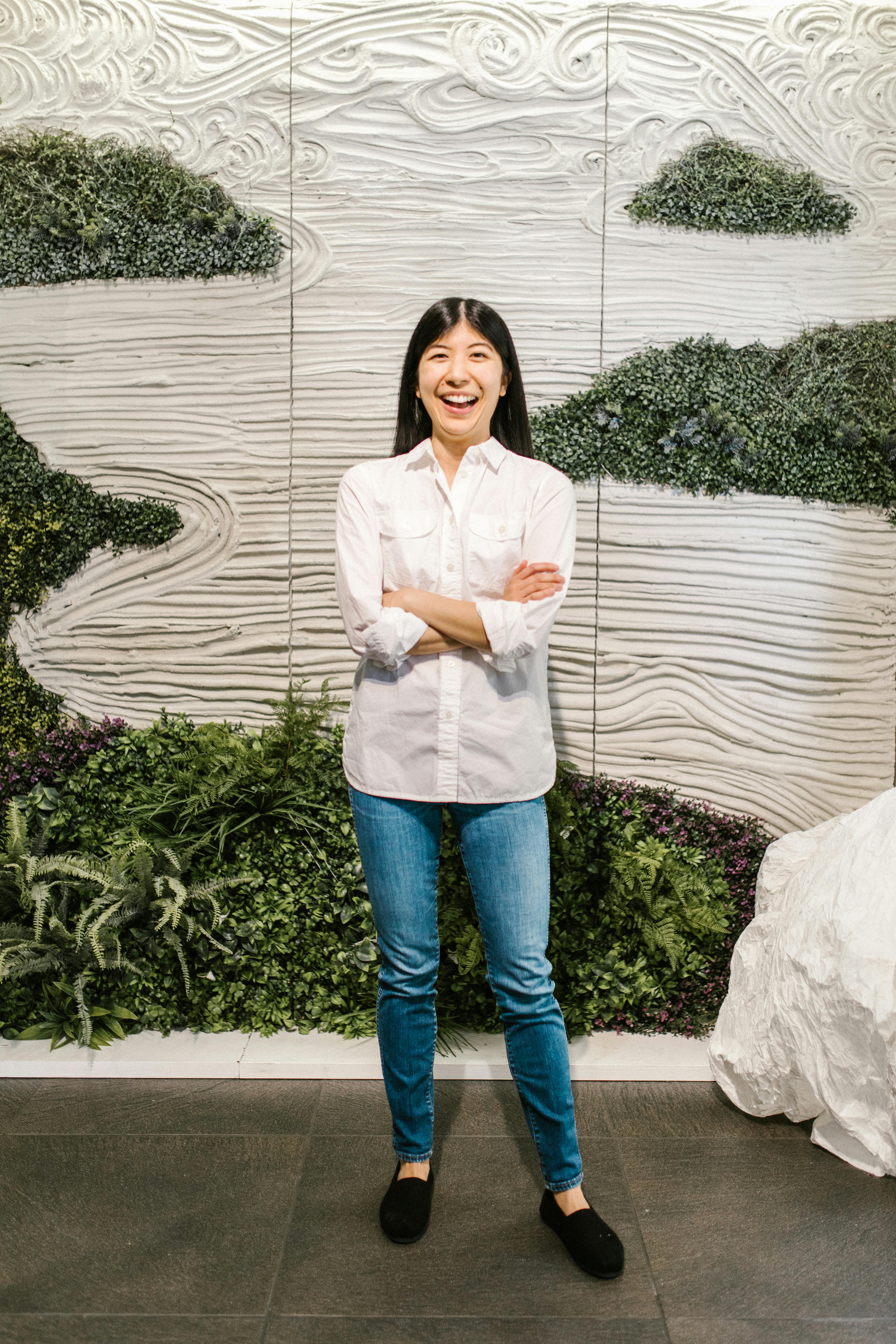 Smiling woman in casual white blouse and jeans, standing against greenery backdrop indoors.