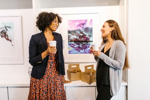 Free Two professional women smiling and chatting during a coffee break in an office art gallery. Stock Photo