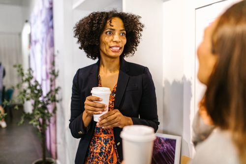 Free African American woman with curly hair holding coffee cup, talking indoors. Warm, lively atmosphere. Stock Photo