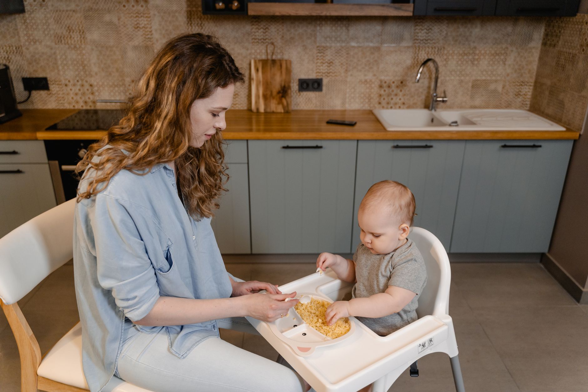 Mother feeds her baby in a high chair in a cozy kitchen, capturing a tender moment in motherhood.