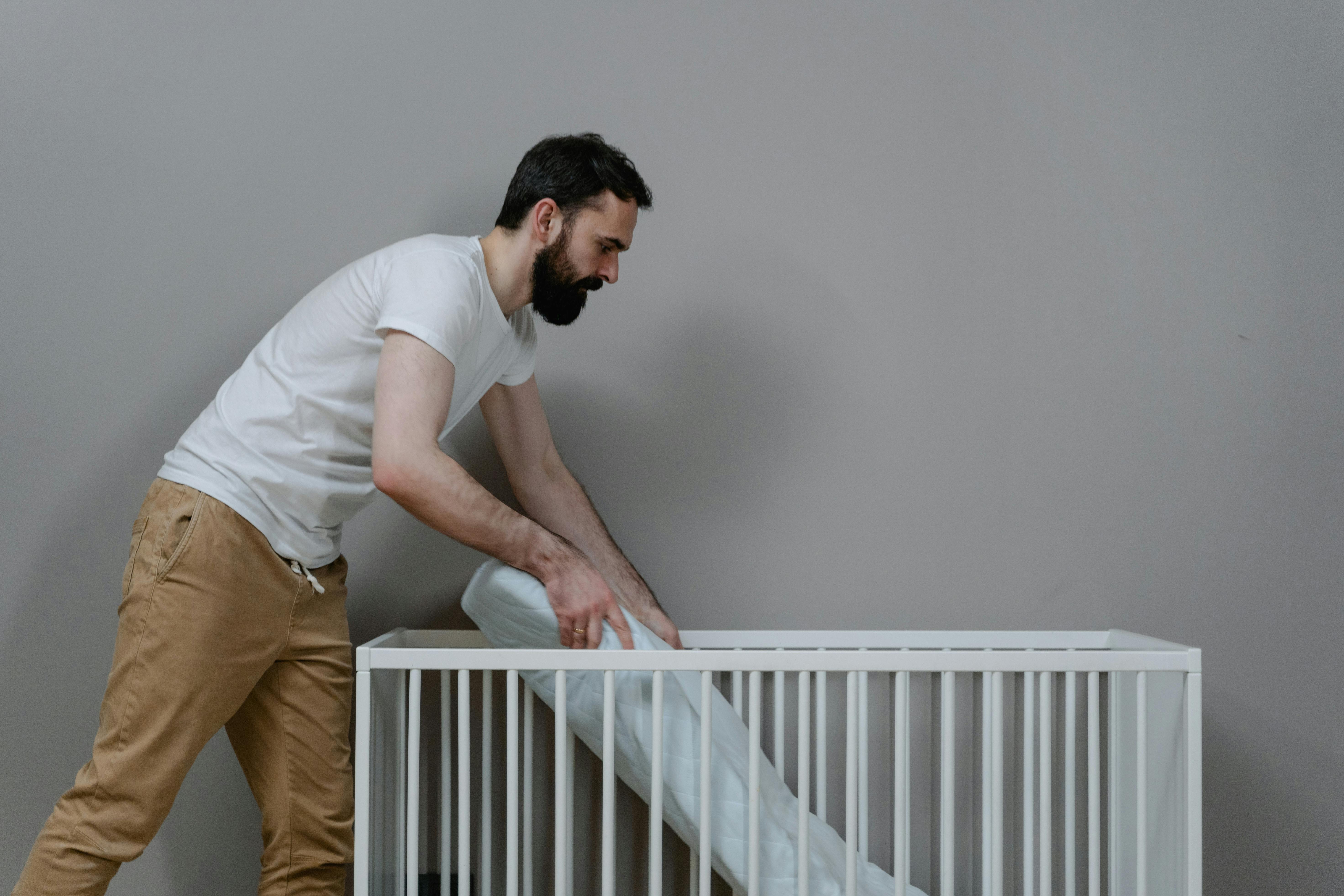 A bearded man setting up a crib in a minimalistic nursery room.