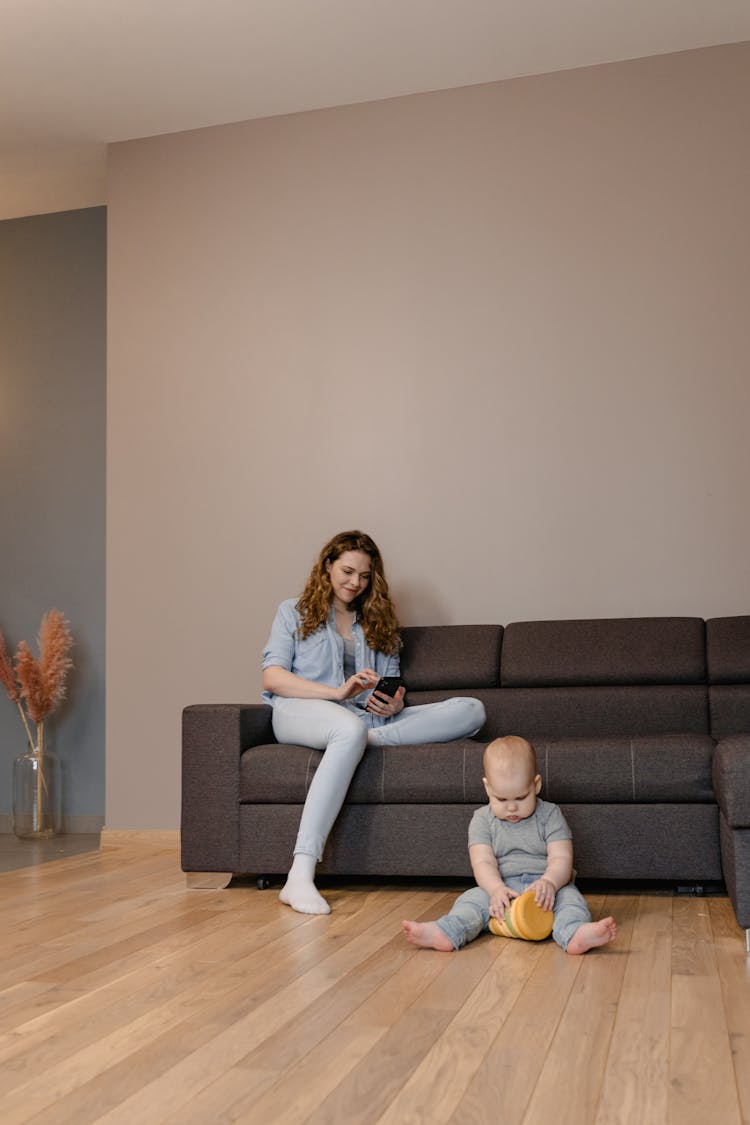 A Woman Sitting On The Couch Using Her Phone While Her Baby Is Sitting On The Floor