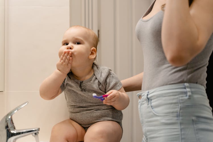 A Cute Baby In Gray Onesie Holding A Toothbrush While Covering Mouth