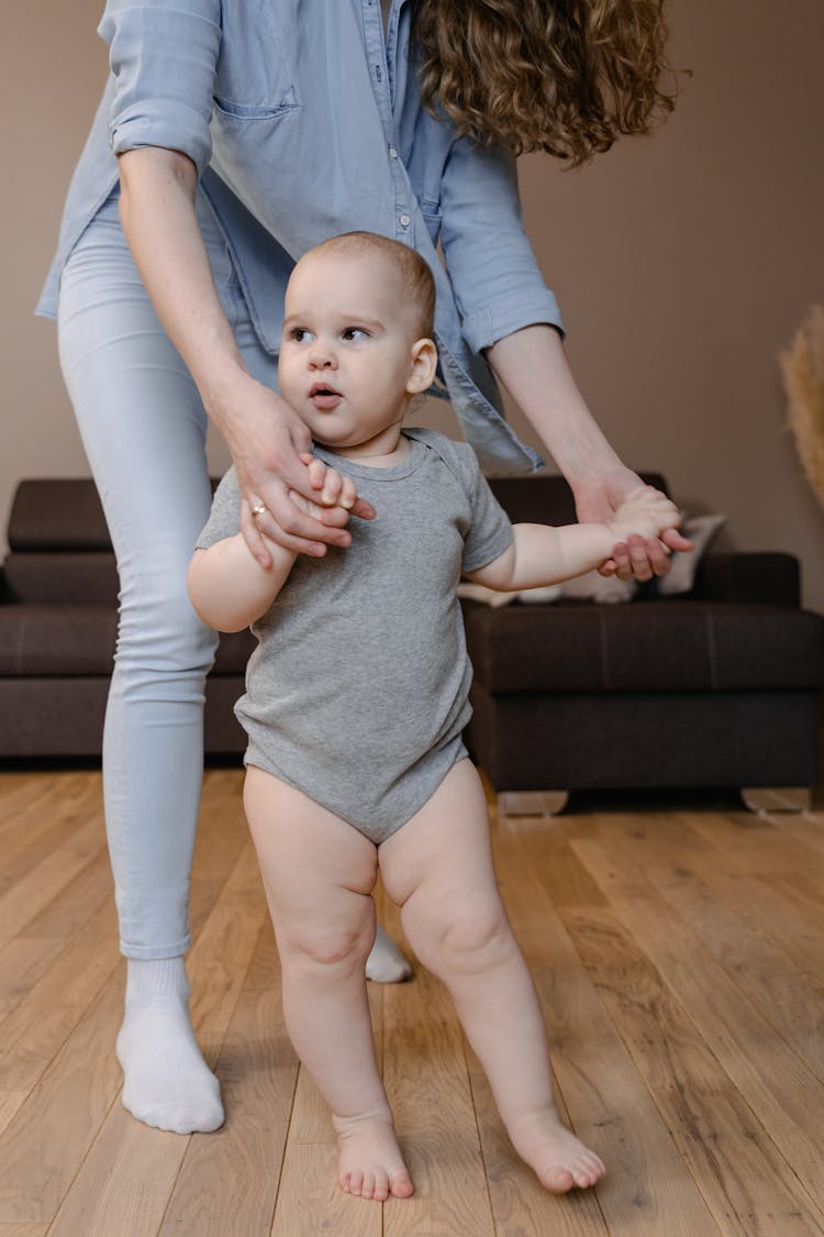 A Cute Baby In Gray Onesie Standing While Holding Hands Of Her Mother