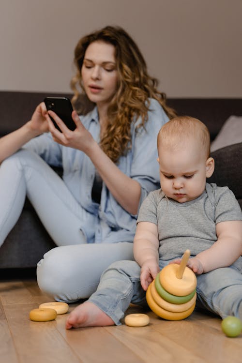 Free A Woman Using Her Mobile Phone while Sitting on the Floor Beside Her Baby Stock Photo
