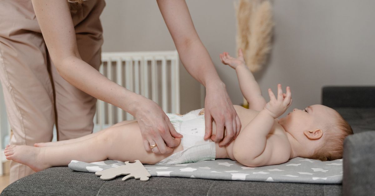 A parent's hands changing a smiling baby's diaper on a gray star-print changing mat, a white crib and pampas grass behind