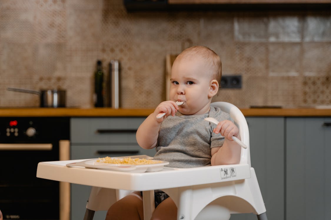 Free A Cut Baby in Gray Shirt Sitting on a High Chair while Eating Food Stock Photo