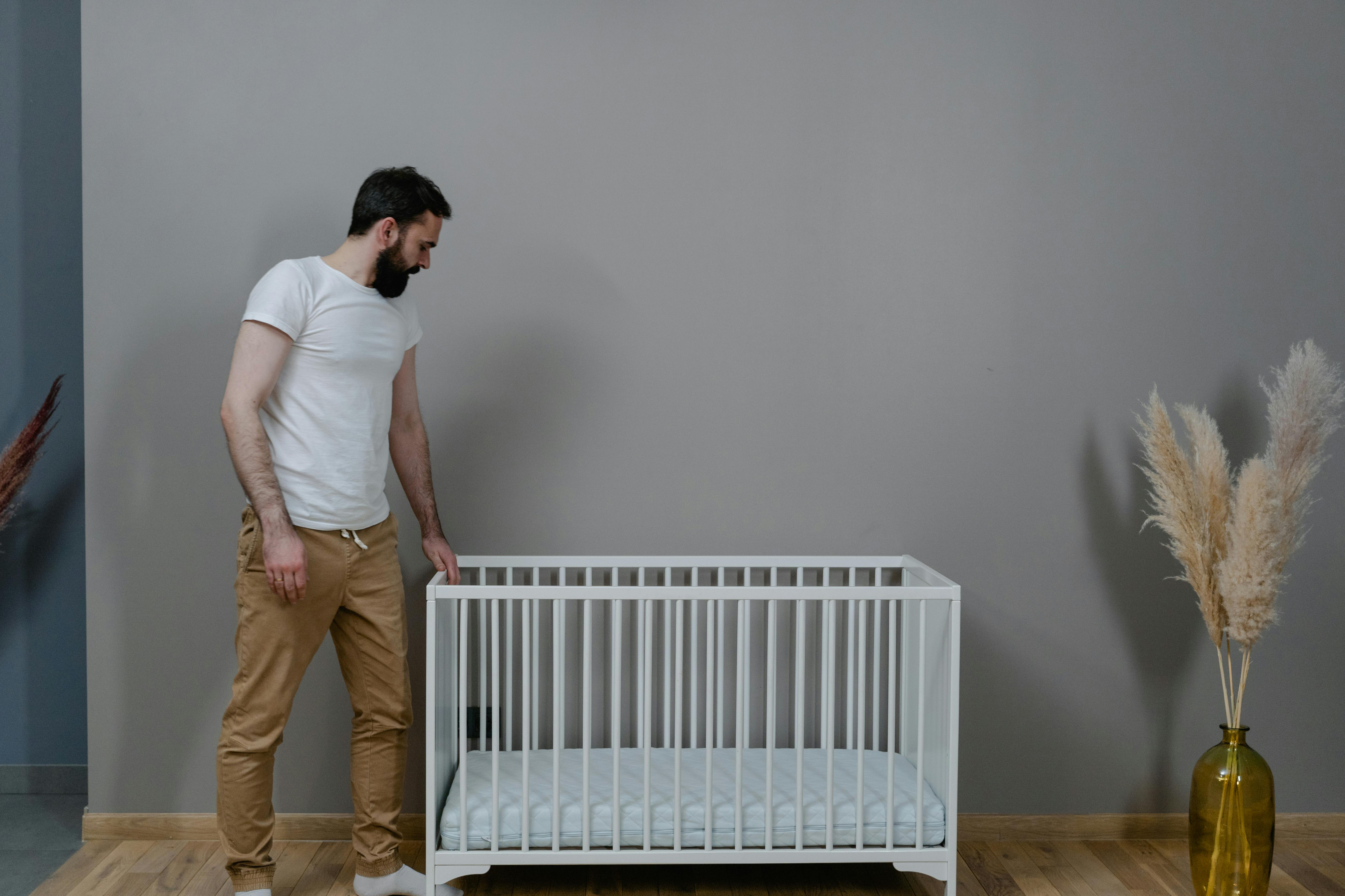 man in white t shirt and brown pants standing beside white wooden crib