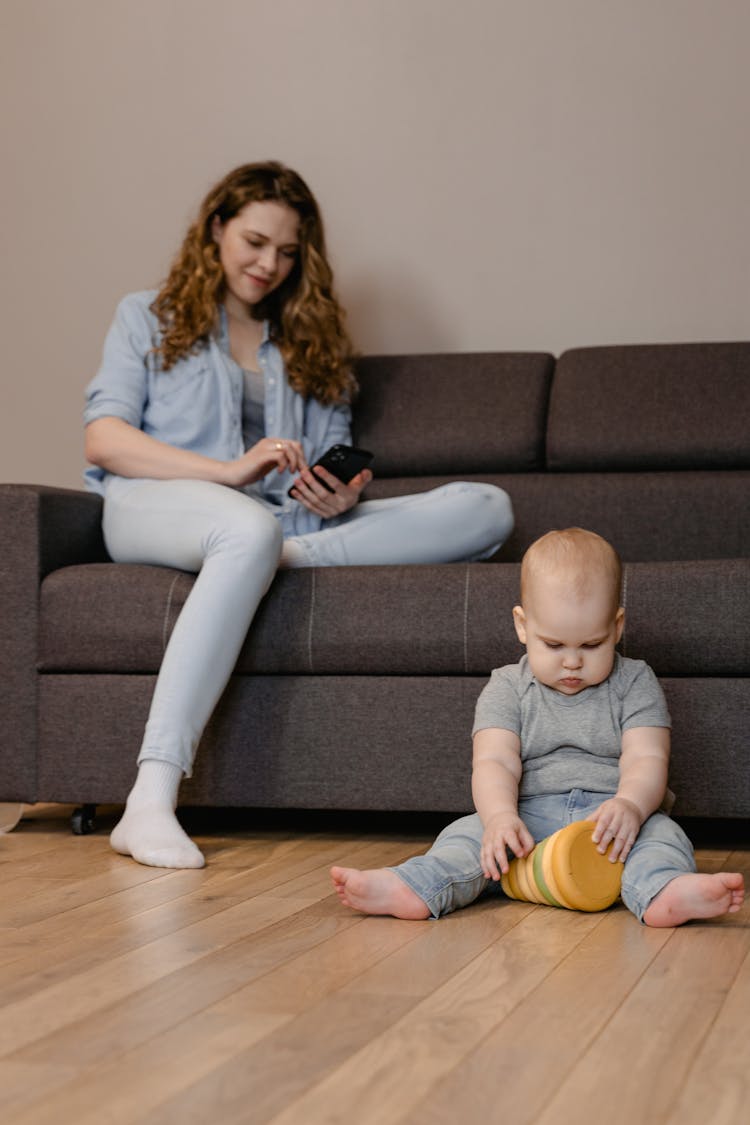 A Woman Using Her Mobile Phone Near Her Baby Sitting On The Floor