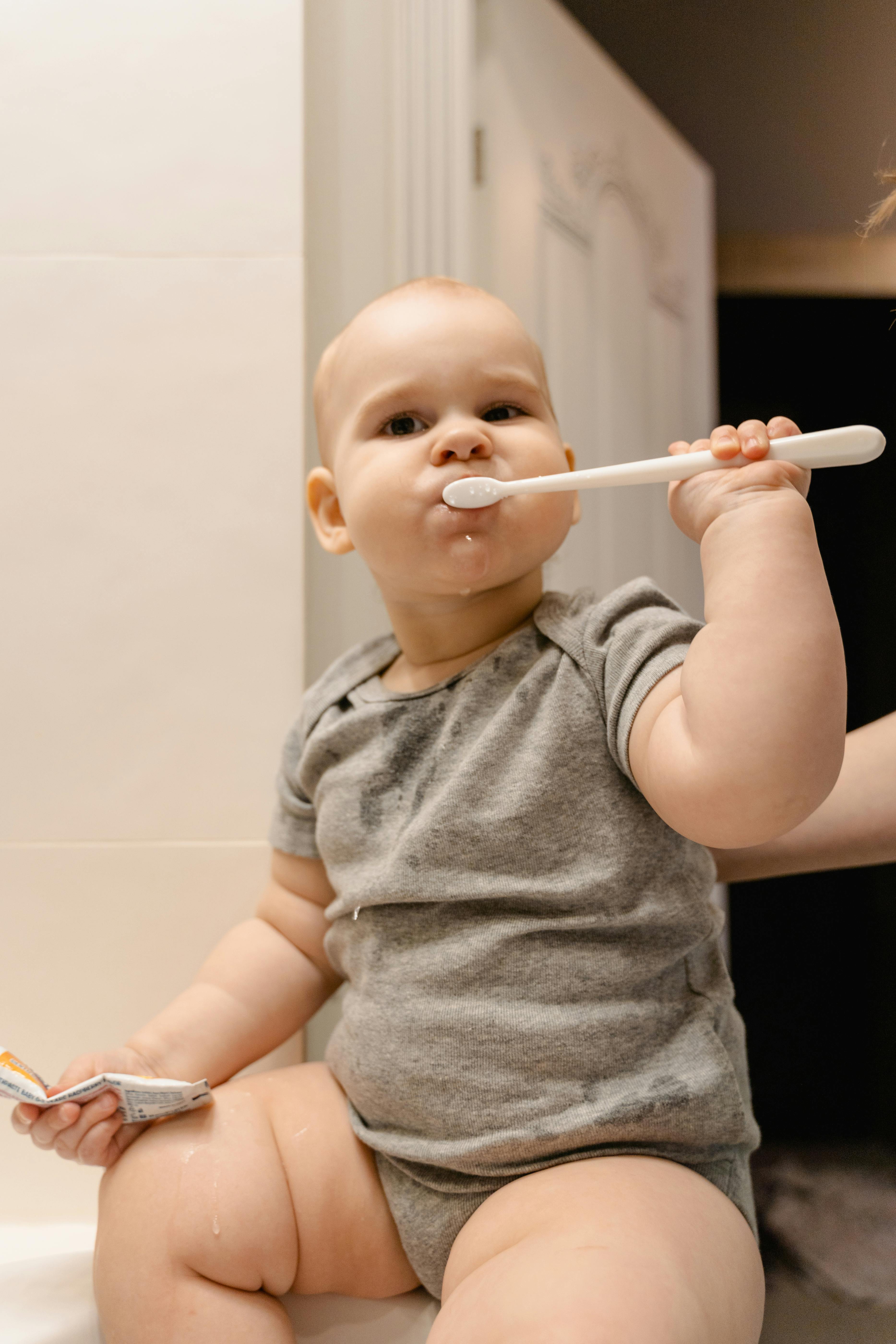Adorable baby in grey outfit brushing teeth, showcasing early hygiene habits.