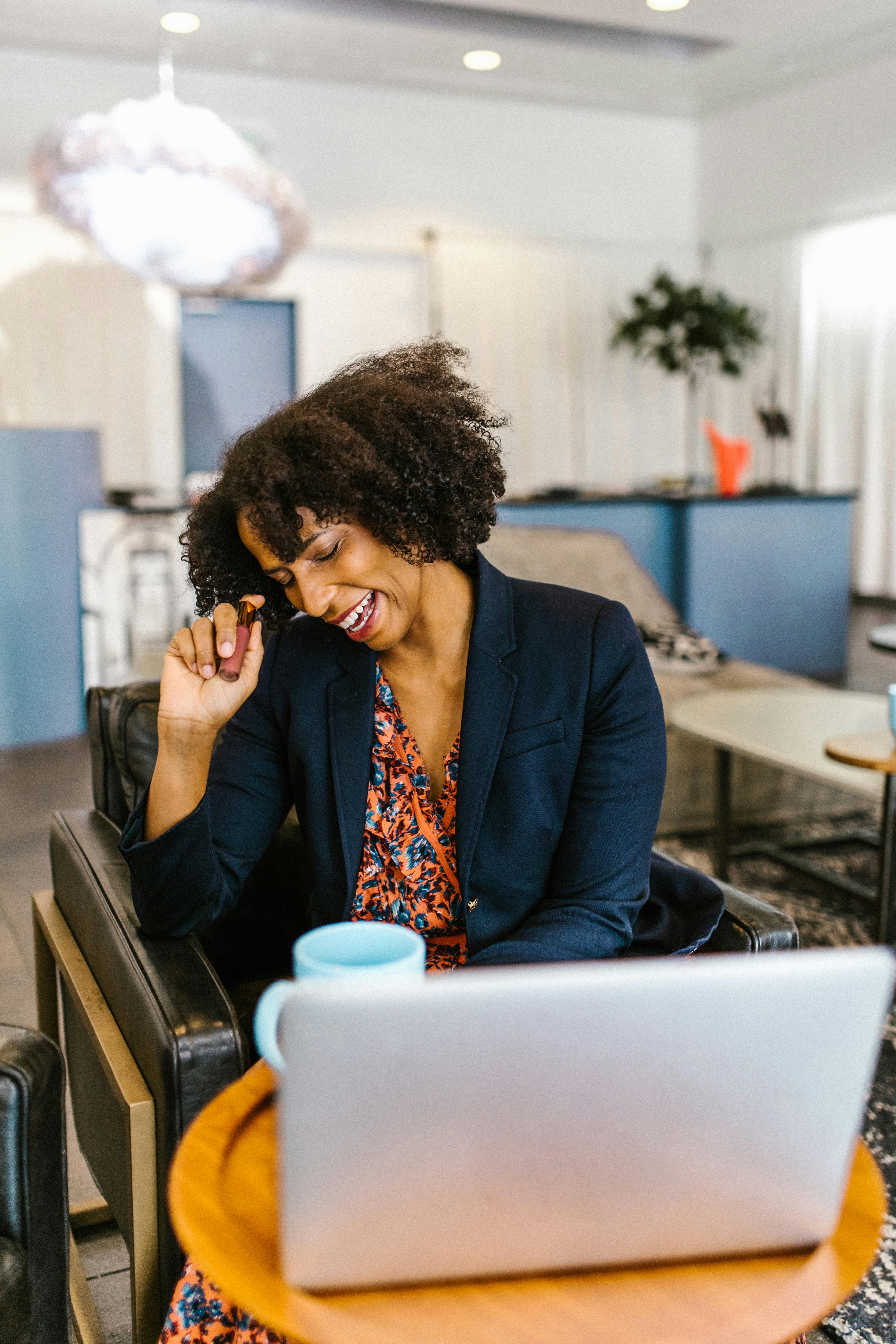 African American woman in office setting, smiling while working on a laptop. Business casual attire.