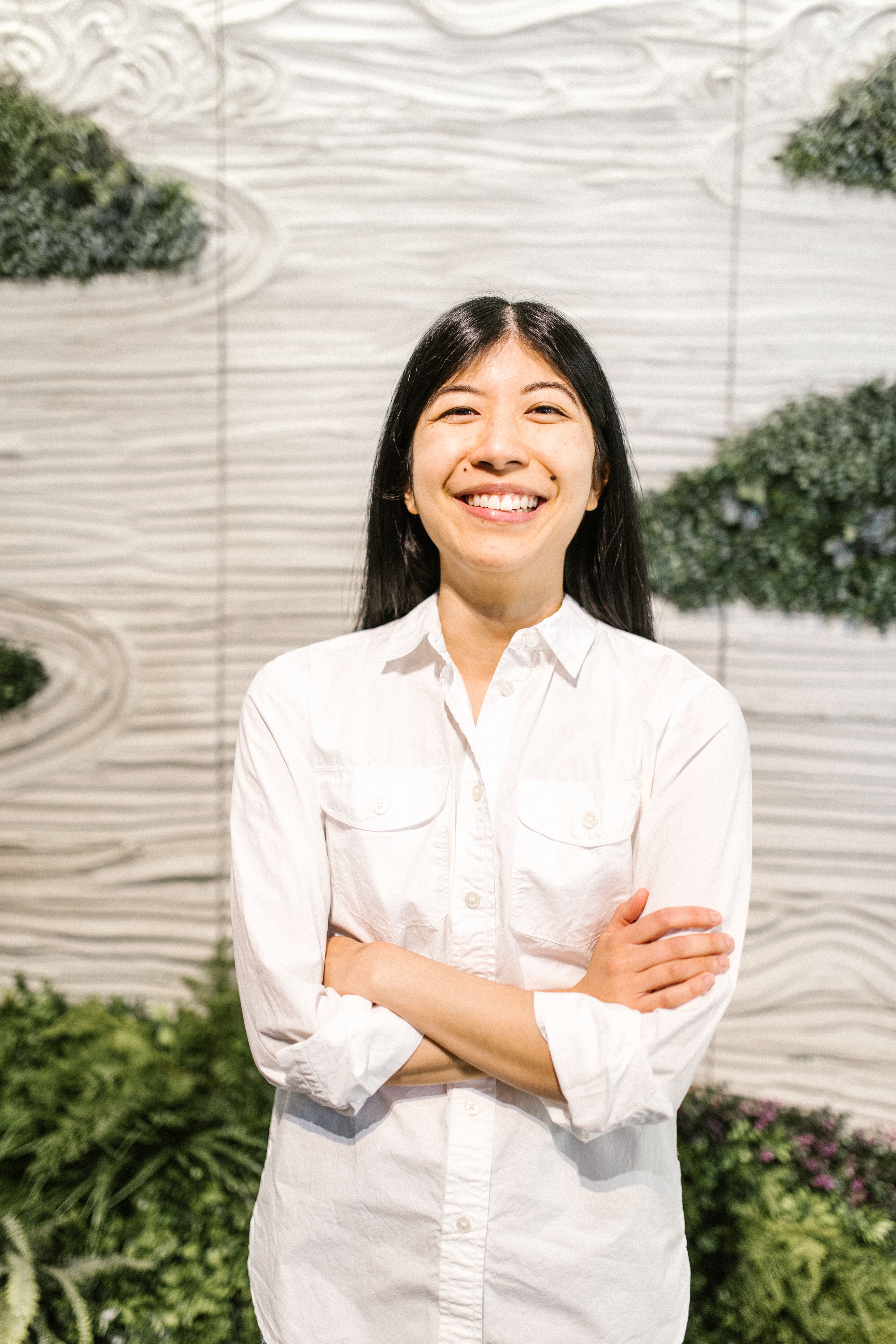 Professional Asian woman with a confident smile, standing in front of a textured wall indoors.