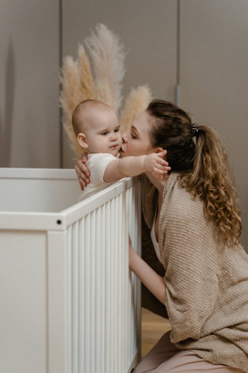 A Woman in Knitted Sweater Kissing Her Baby on the Crib